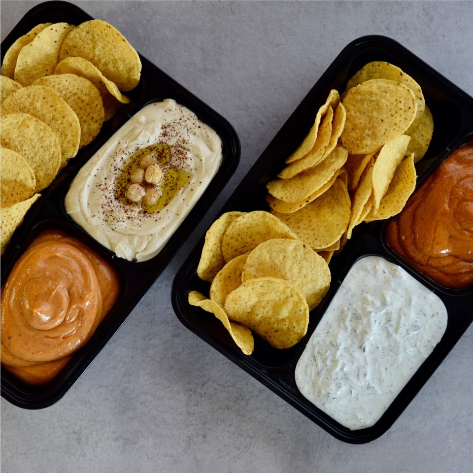 Two black trays filled with tortilla chips, hummus, and two other dips.