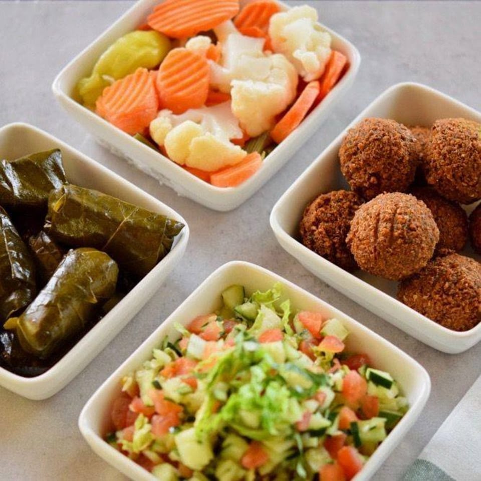Four white square dishes filled with Mediterranean falafel, salad, pickled vegetables, and stuffed grape leaves.